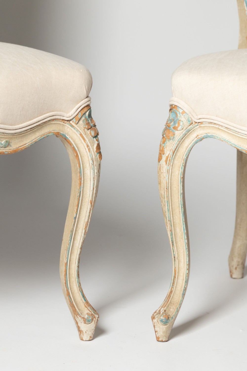 A pair of antique French side chairs featuring distressed off-white painted frames with floral crest carvings, cabriole legs, and neutral linen upholstery. View IX