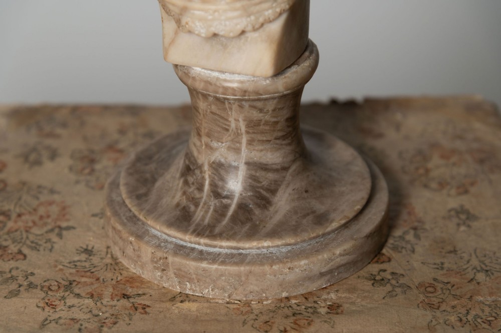 A carved cream alabaster bust depicting a female figure with stylized hair atop a polished turned stone socle. View V