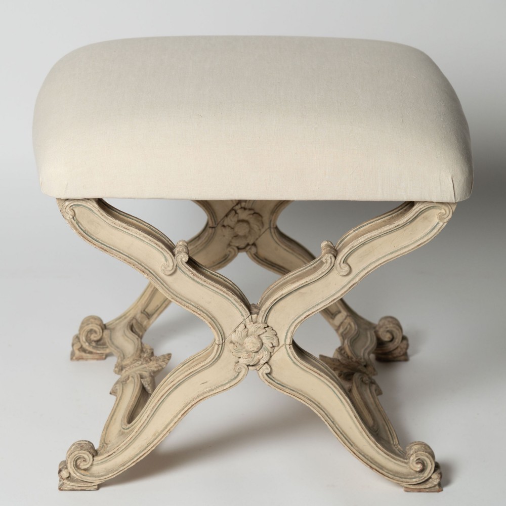 An antique French X-frame stool featuring a painted cream carved wooden base with rosette detailing and a neutral linen upholstered cushion.