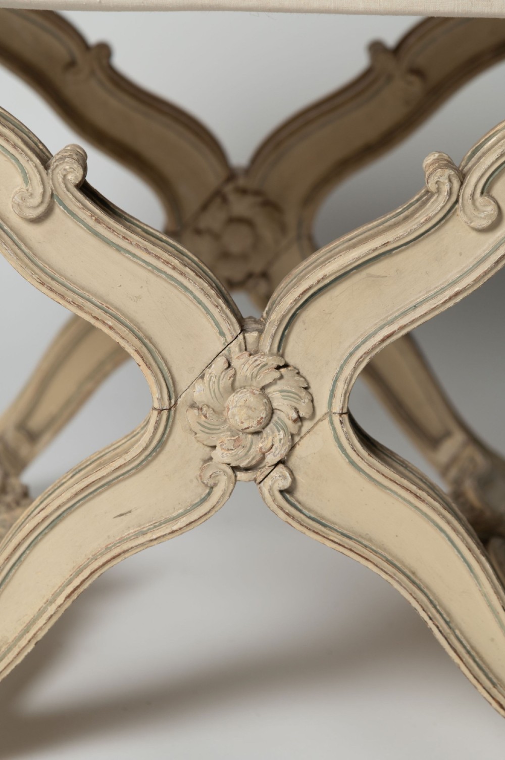 An antique French X-frame stool featuring a painted cream carved wooden base with rosette detailing and a neutral linen upholstered cushion. View III