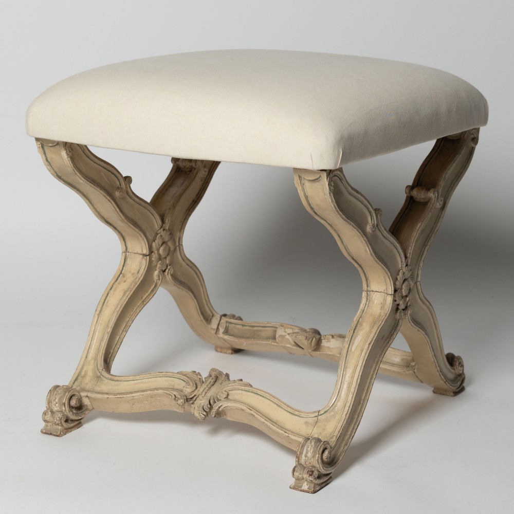 An antique French X-frame stool featuring a painted cream carved wooden base with rosette detailing and a neutral linen upholstered cushion. View V