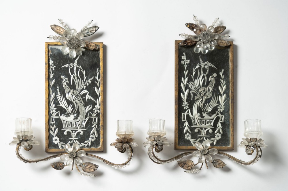 A pair of French gilt metal wall sconces featuring verre eglomise panels depicting birds, with floral crystal cresting and two scroll arms.