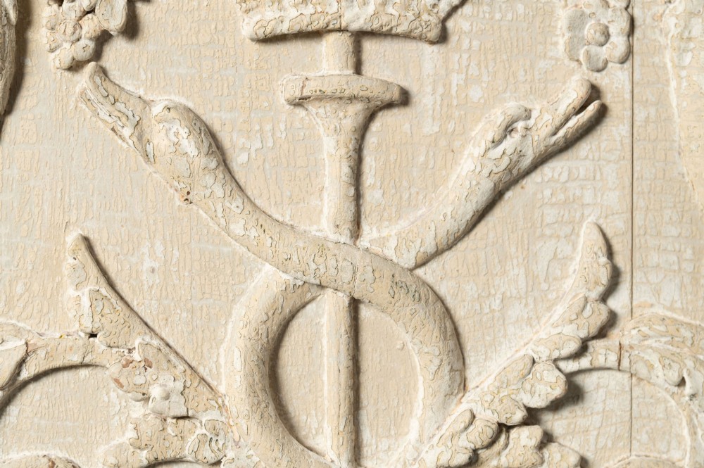 A rectangular off-white painted wood panel featuring an intricate raised relief carving of heraldic cranes, a fruit-filled urn, caduceus serpents, and laurel swags within a moulded frame. View V