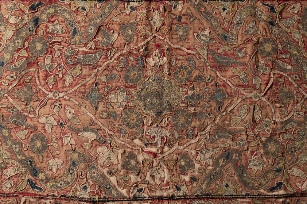 A rectangular 18th-century hand-embroidered textile featuring a central diamond medallion with floral arabesques and border patterns in shades of muted pink, sage green, and cream. View III