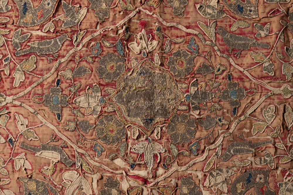 A rectangular 18th-century hand-embroidered textile featuring a central diamond medallion with floral arabesques and border patterns in shades of muted pink, sage green, and cream. View IV
