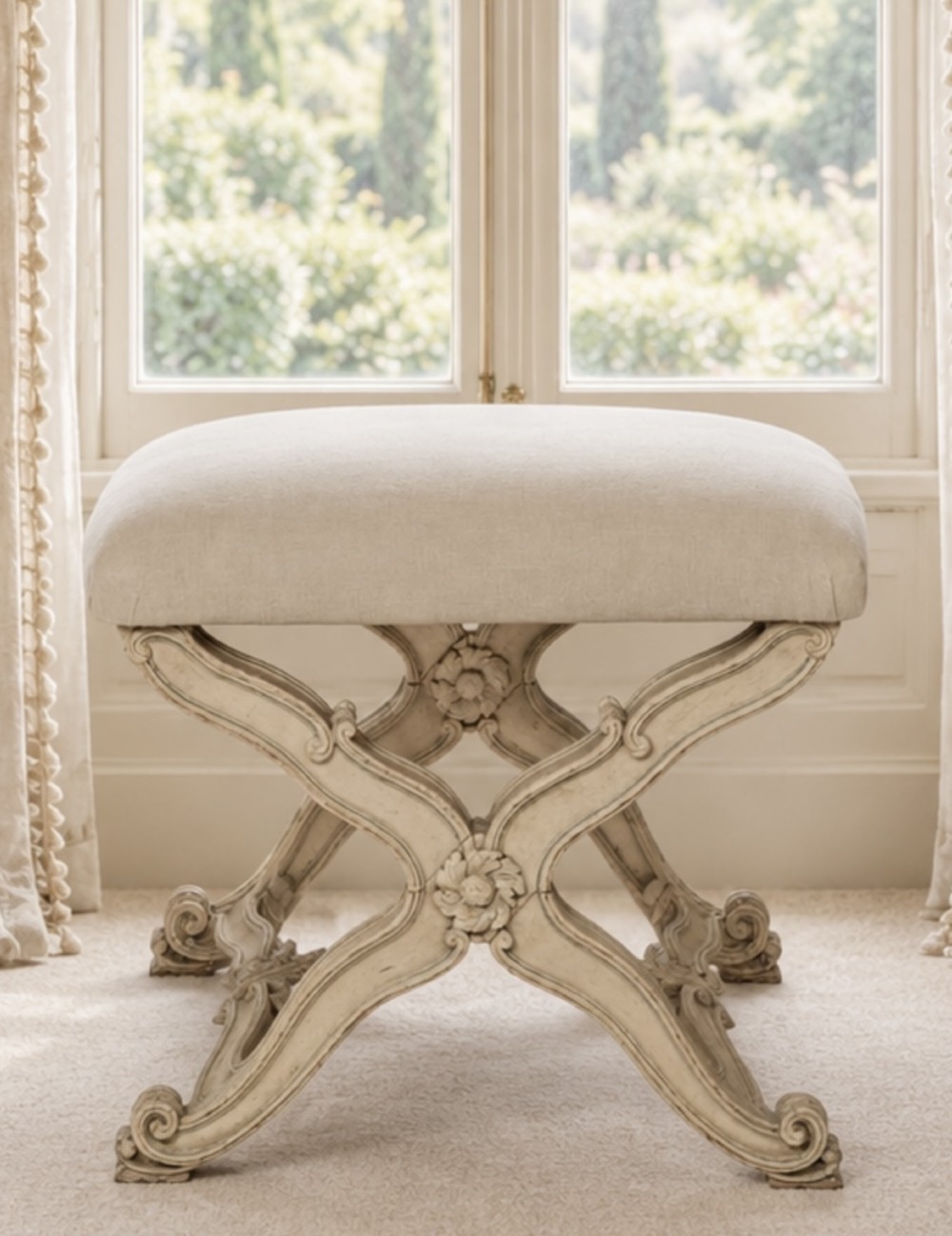 An antique French X-frame stool featuring a painted cream carved wooden base with rosette detailing and a neutral linen upholstered cushion.