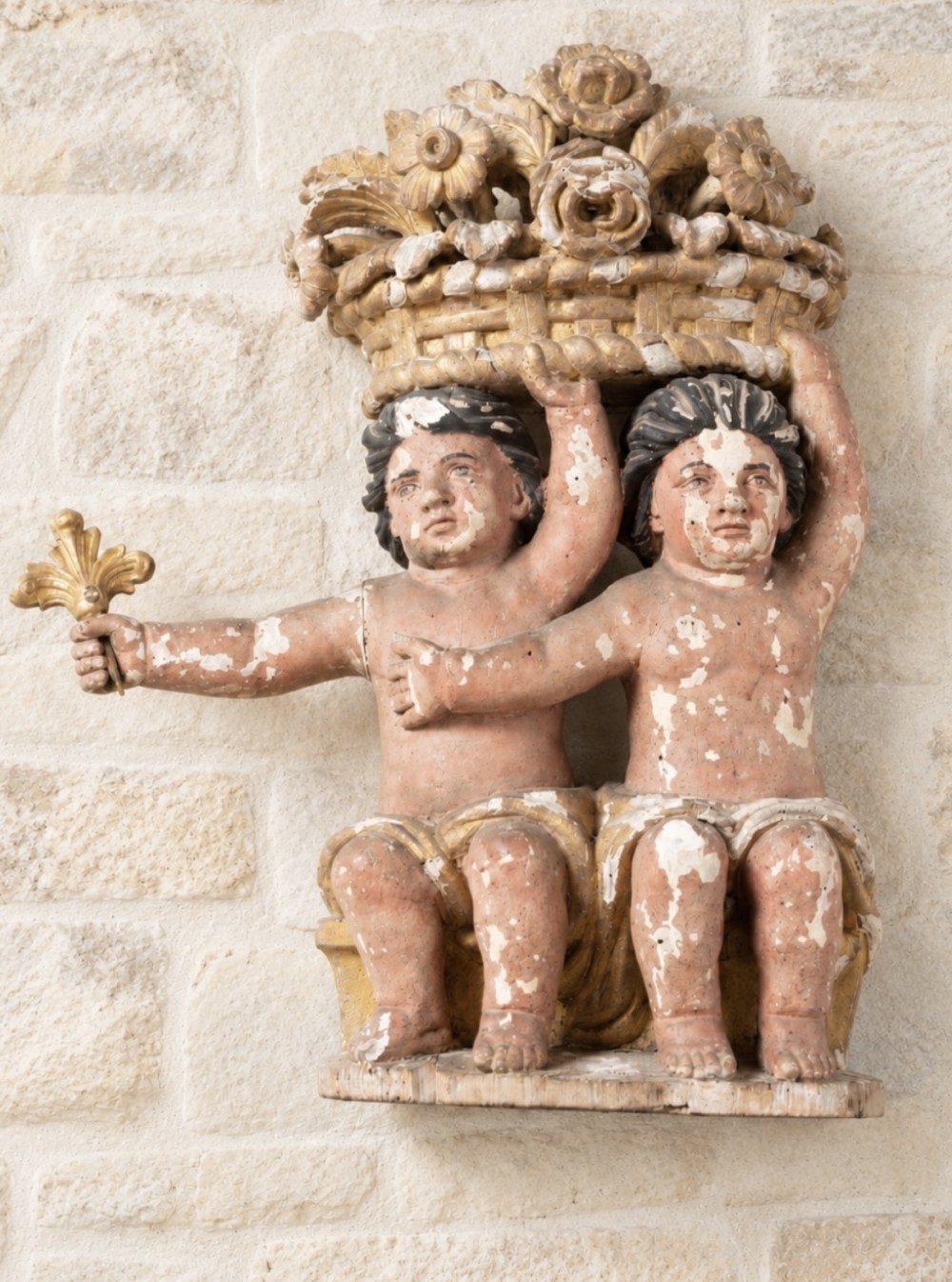 A carved wood polychrome and gilded relief sculpture featuring two seated putti supporting an elaborate basket of flowers on their heads.