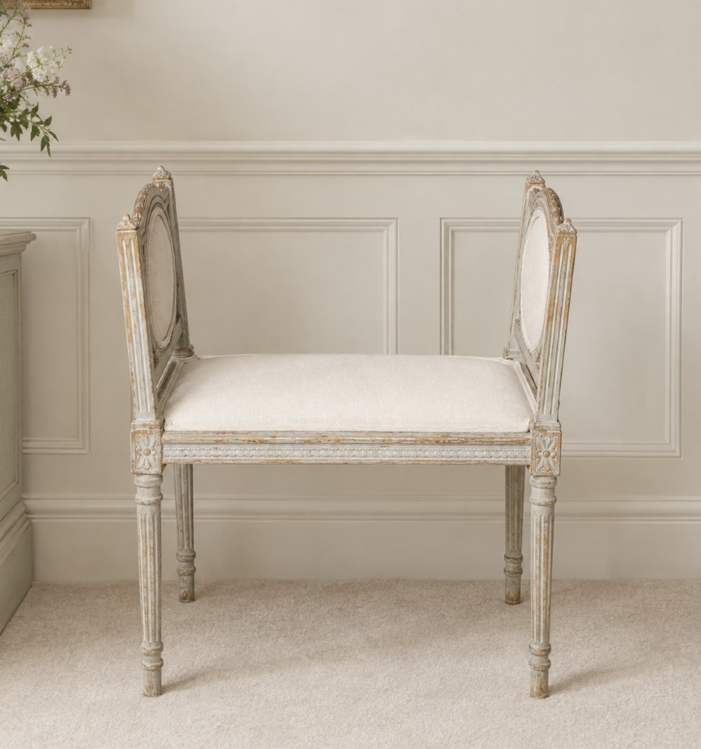 A Louis XVI style antique French upholstered stool featuring fluted legs, carved oval side handles, and a distressed grey painted finish with off-white linen seating.