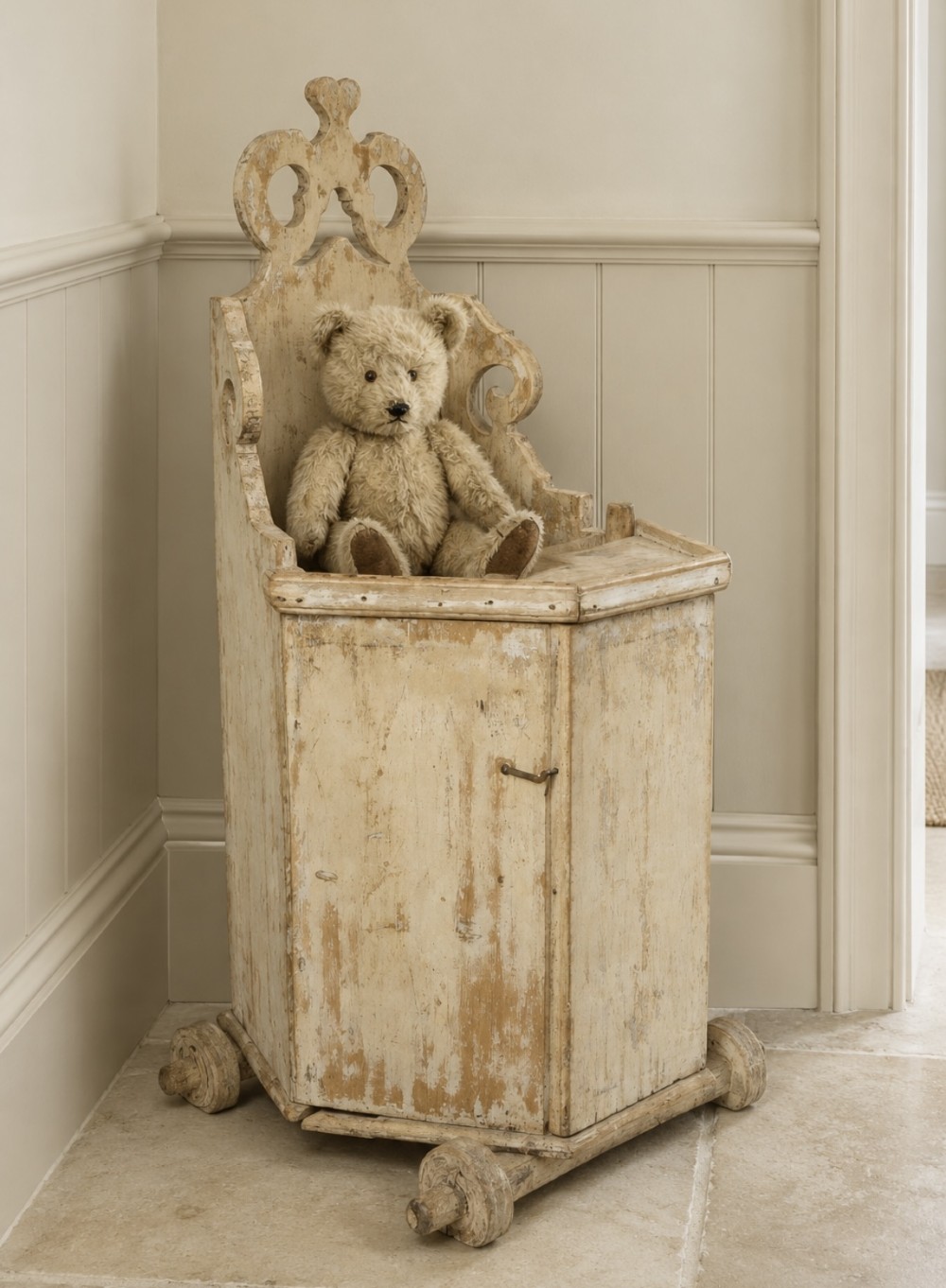 A distressed cream painted pine Swedish antique highchair featuring a carved cresting backrest and wooden wheels.