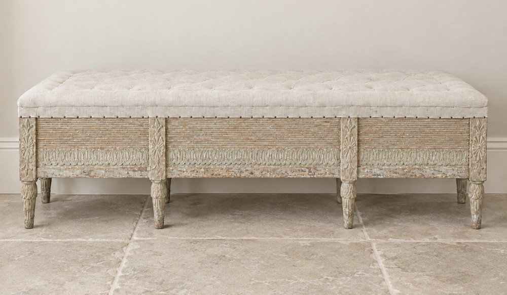 An upholstered long wooden bench featuring a button-tufted linen top, distressed pale grey paintwork, six fluted legs, and relief-carved floral motifs.