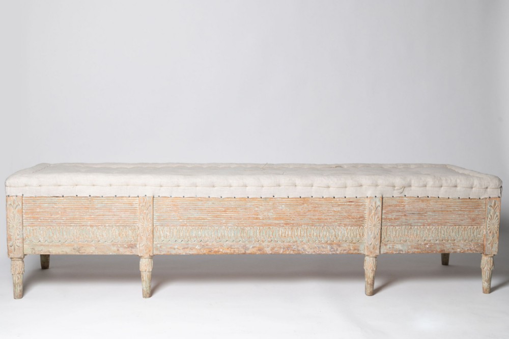 An antique Swedish long stool featuring a distressed painted pine frame with eight fluted tapered legs and a neutral linen upholstered top.