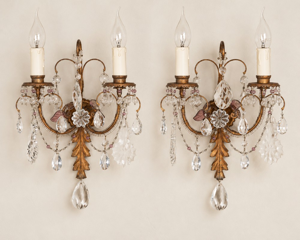 A pair of two-arm gilt metal wall lights decorated with crystal teardrop pendants, glass rosettes, and candle-shaped light sleeves.