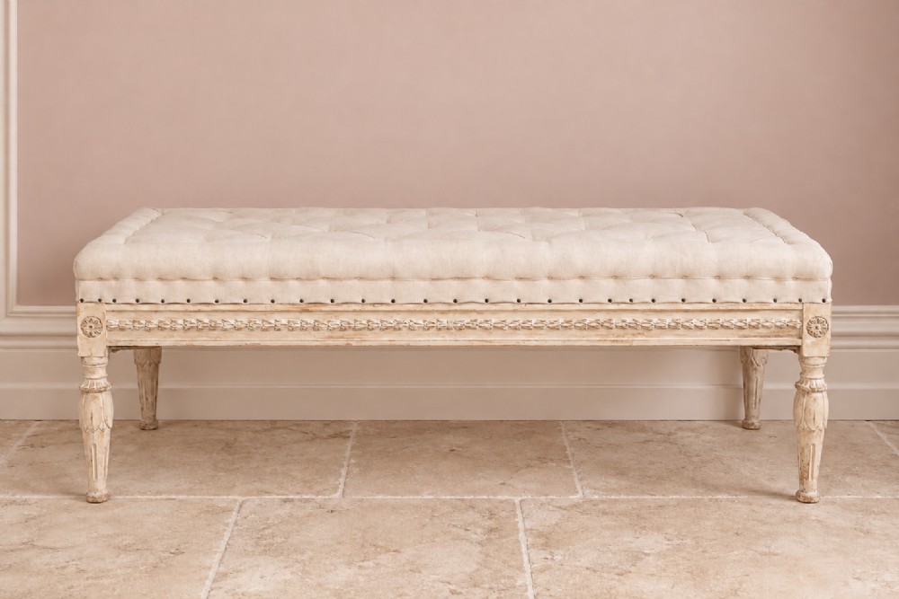 An off-white painted Gustavian style long bench featuring button-tufted linen upholstery, carved floral frieze, and fluted legs.