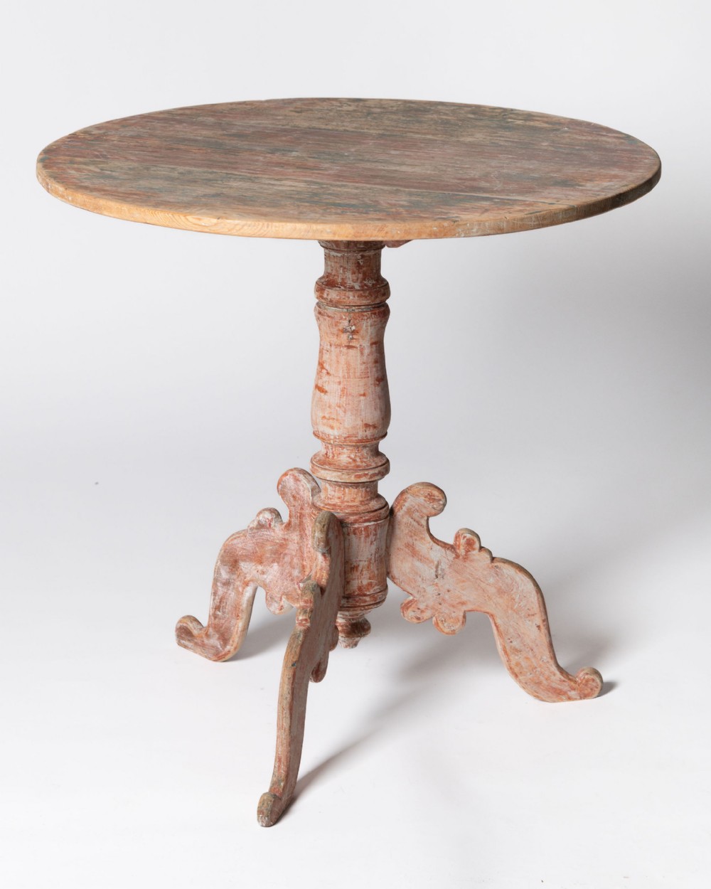 An antique Swedish tilt-top table featuring a round pine surface, a turned central pedestal, and a carved tripod base with faded terracotta paint.