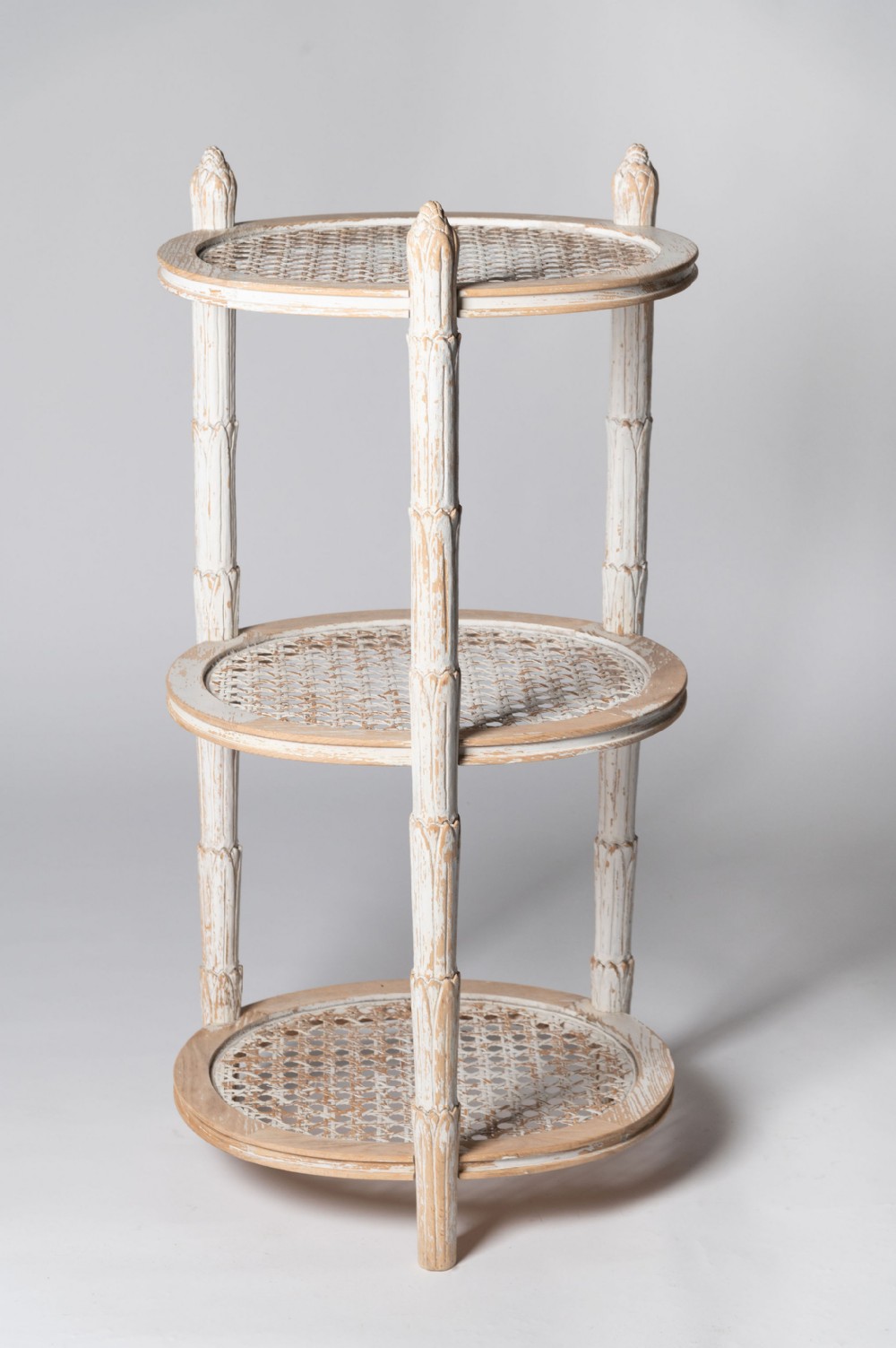 A three-tiered circular French etagere with distressed off-white painted woodwork and woven cane shelf surfaces.
