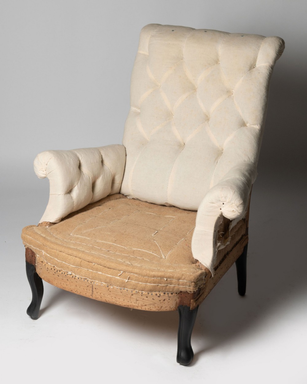 An antique French armchair upholstered in cream calico with deep buttoning across the backrest and ebonised cabriole front legs.