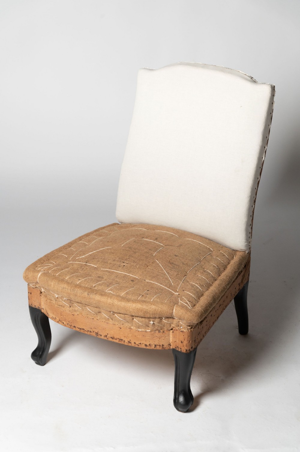 An antique French armless slipper chair with a hessian upholstered seat, calico backrest, and ebonised cabriole front legs.