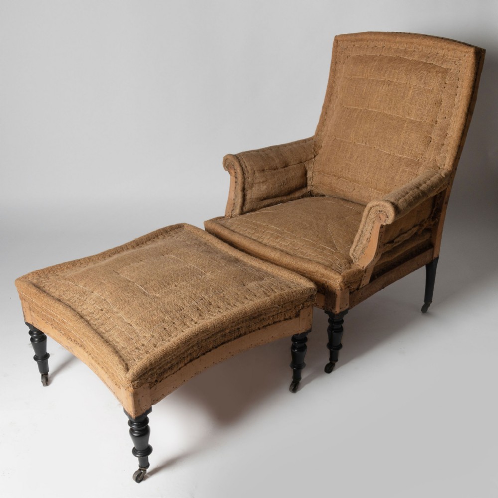 An antique French armchair and matching footstool covered in coarse beige hessian upholstery with black turned wooden legs on castors.