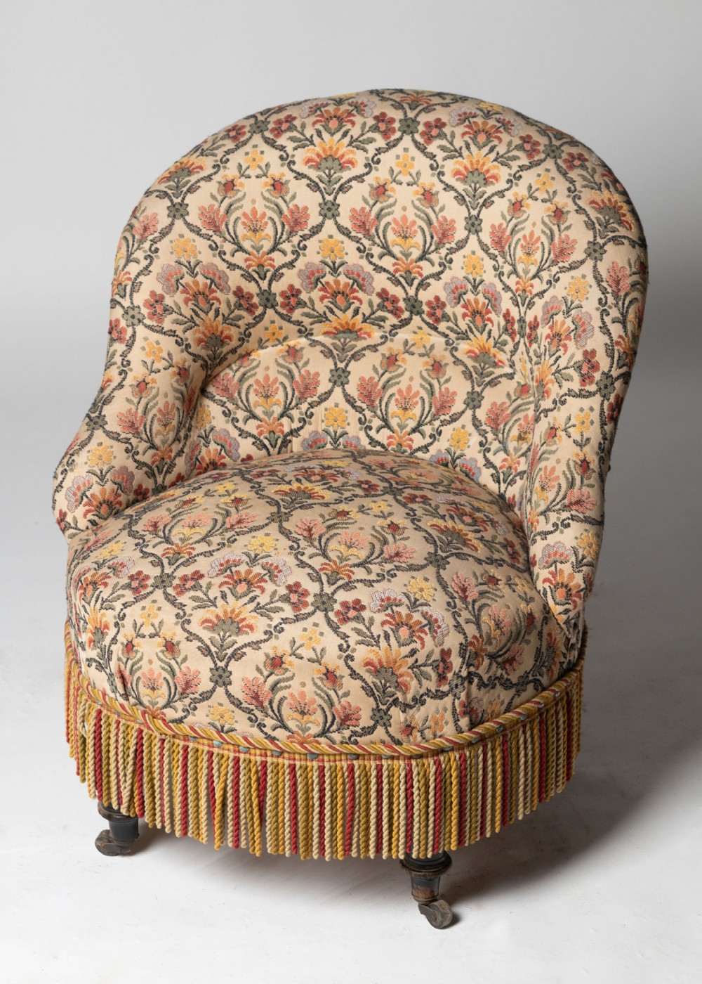 A low-slung French crapaud armchair upholstered in a floral tapestry fabric with a fringe trim base and turned legs on castors.