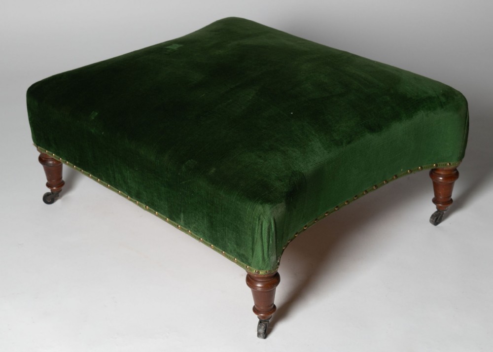 A large square antique French footstool upholstered in dark green velvet with brass stud trim and turned wooden legs on castors.