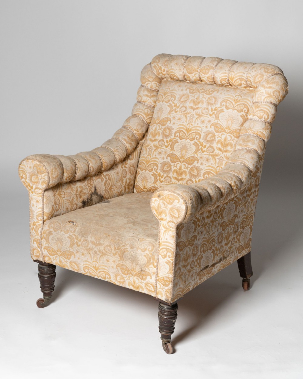 An antique Victorian upholstered armchair with gold floral damask fabric, deep channelled border trim, turned dark wooden legs, and brass castors.