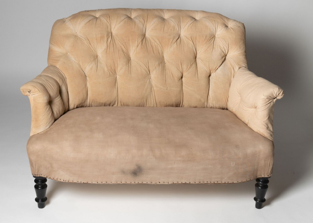 A small antique French sofa featuring deep button-tufted beige velvet upholstery, padded armrests, and four dark turned wooden legs.