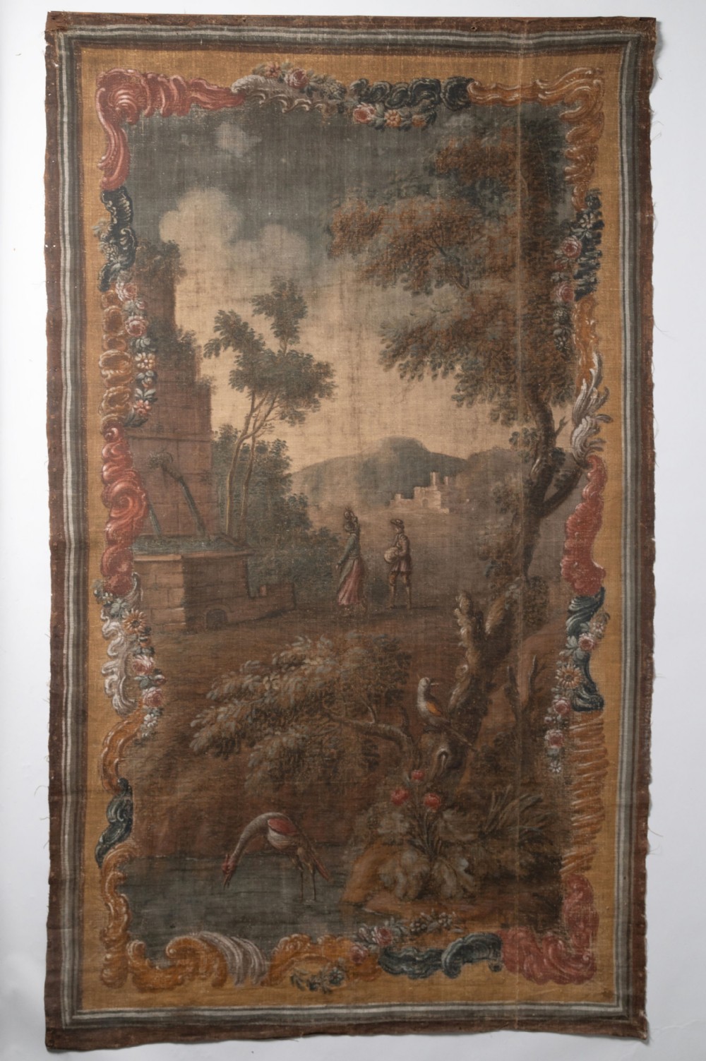 An antique vertical French textile tapestry featuring a pastoral landscape scene with figures, foliage, and water enclosed within a decorative scrolling border.