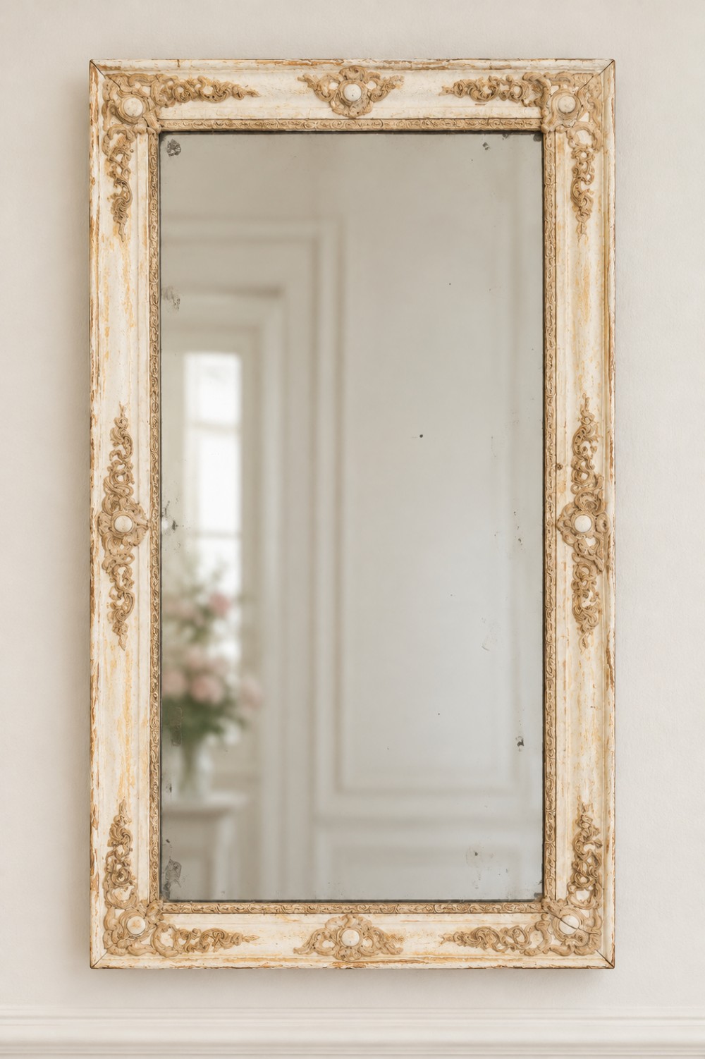 A tall rectangular antique French wall mirror featuring a distressed cream frame decorated with carved foliate mouldings and foxed mirror glass.