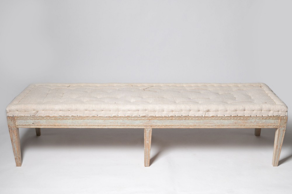An antique Swedish Gustavian long stool featuring a pale grey painted frame with tapered fluted legs, carved corner rosettes, and a button-tufted cream linen seat.