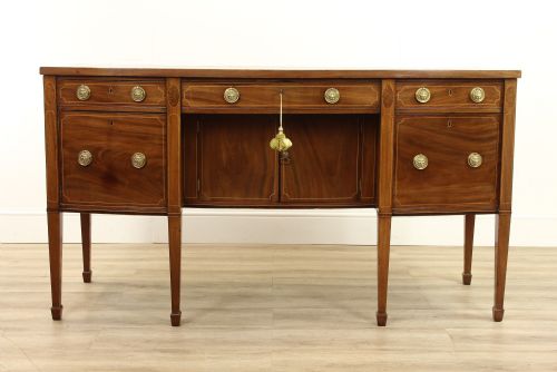 fig 1. A Georgian mahogany bow-fronted sideboard with brass lion mask ring handles, inlay stringing, and tapered square legs on a pale floor.