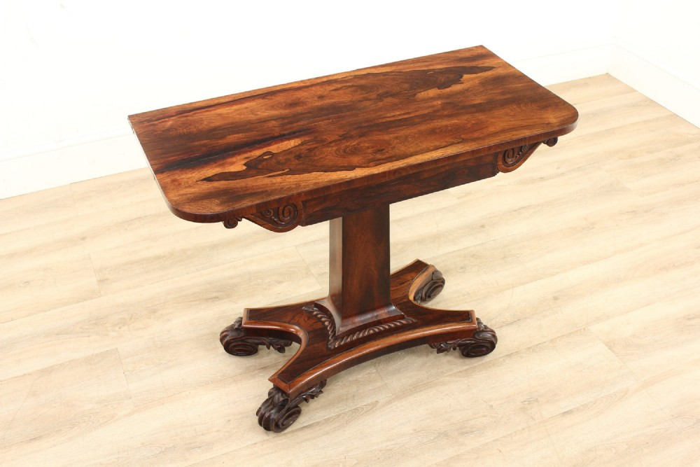 A William IV rosewood pedestal console table featuring a figured rectangular top, carved scroll frieze brackets, a central column, and a quadruped platform base with scroll feet.