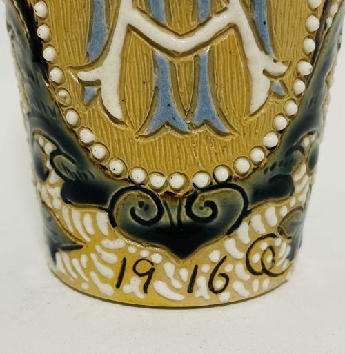 fig 1. An Arts and Crafts salt-glazed stoneware beaker from 1916 featuring a central monogram framed by white beaded slip, blue detailing, and an ochre textured background.