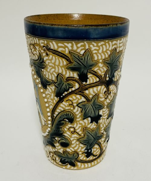 fig 2. An Arts and Crafts salt-glazed stoneware beaker from 1916 featuring a central monogram framed by white beaded slip, blue detailing, and an ochre textured background.