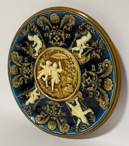 fig 1. A circular Victorian majolica charger featuring relief figures of children and scrollwork in cobalt blue, ochre, and ivory glazes.