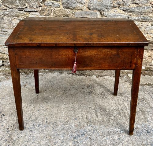 fig 1. A Georgian solid oak clerks desk featuring a hinged sloped writing top, a key with tassel inserted into the central keyhole, and four square tapered legs.