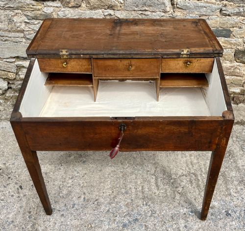 fig 2. A Georgian solid oak clerks desk featuring a hinged sloped writing top, a key with tassel inserted into the central keyhole, and four square tapered legs.