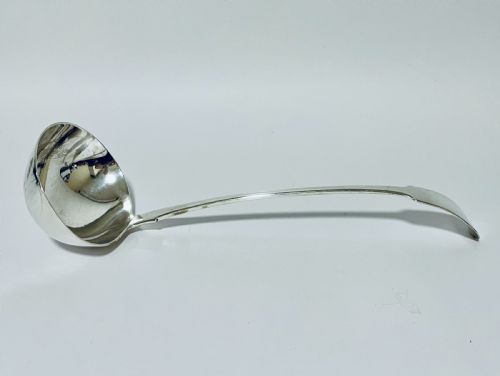 fig 1. A polished Georgian silver serving ladle featuring a deep circular bowl and a long curved handle resting on a plain light surface.