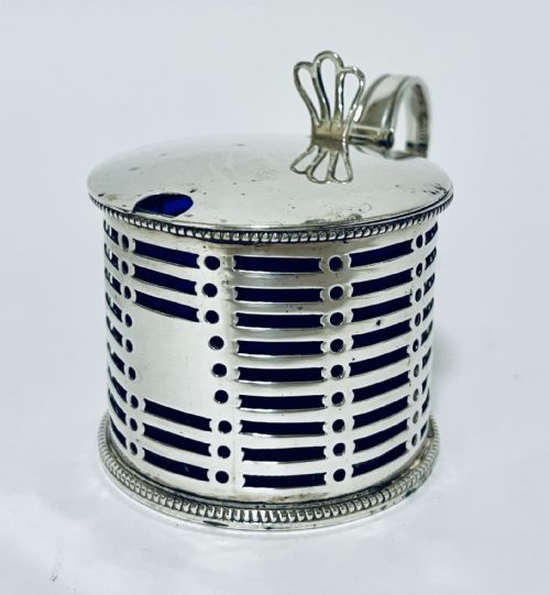 fig 1. A cylindrical pierced silver mustard pot featuring a hinged lid, side handle, beaded borders, and a cobalt blue glass liner.
