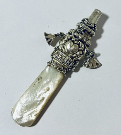 fig 1. An antique silver baby rattle and teether featuring a flat mother of pearl handle, suspended silver bells, an integrated whistle top, and embossed lettering spelling DARLING.