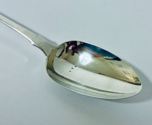 fig 1. A Georgian sterling silver serving spoon featuring a polished oval bowl and a classic fiddle pattern handle resting against a plain light background.