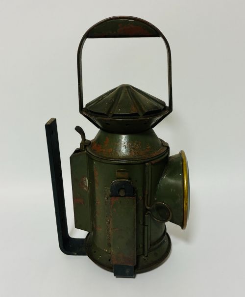 fig 1. A vintage olive green painted metal railway signal lamp featuring a large round glass lens with a brass rim and a tall arched carrying handle.