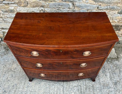 fig 1. An antique Regency mahogany chest of drawers featuring a curved bowfront shape, three drawers with oval brass handles, and splayed bracket feet.