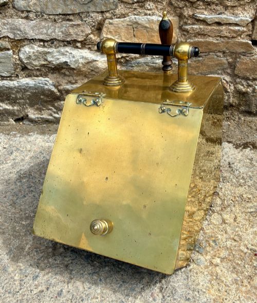 fig 1. An antique brass sloped coal scuttle featuring a turned wooden carrying handle, a hinged lid, and an attached brass shovel at the rear.