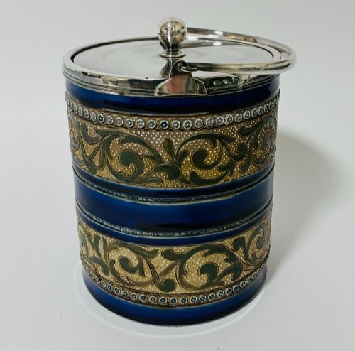 fig 1. A cylindrical Victorian Doulton Lambeth stoneware biscuit barrel featuring cobalt blue and beige scrollwork decoration, fitted with a silver-plated lid, swing handle, and finial.