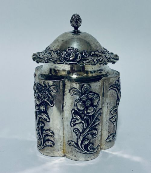 fig 1. A lobed silver tea caddy with embossed floral patterns, a wavy rimmed lid, and a pinecone-shaped finial.