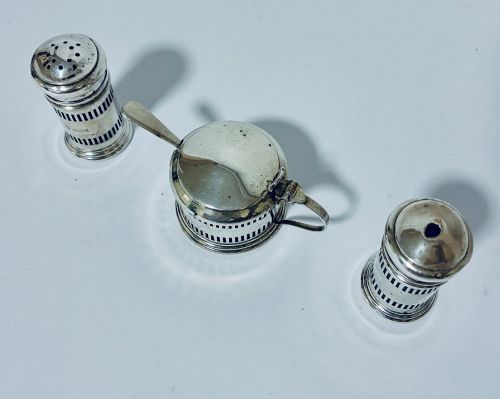 fig 2. An antique three-piece silver cruet set featuring pierced vertical slot details, a mustard pot with lid and spoon, and two matching pepper casters.