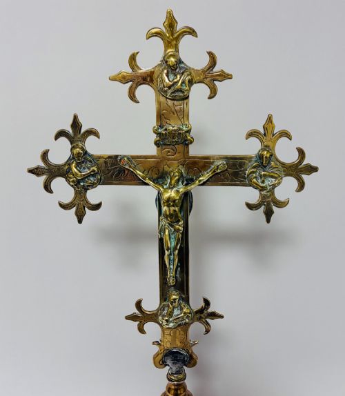 fig 1. An antique brass processional cross featuring a detailed crucifix figure, fleur-de-lis terminal accents, and engraved motifs, mounted on a turned dark wood stand.
