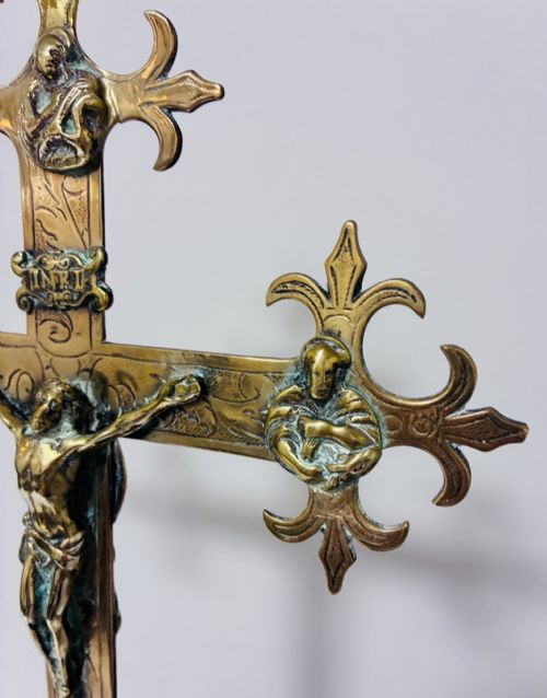 fig 2. An antique brass processional cross featuring a detailed crucifix figure, fleur-de-lis terminal accents, and engraved motifs, mounted on a turned dark wood stand.