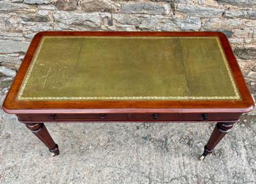 fig 1. A Victorian mahogany writing table featuring a green gilt-tooled leather writing surface, two drawers with turned knob handles, and turned tapered legs resting on ceramic castors.