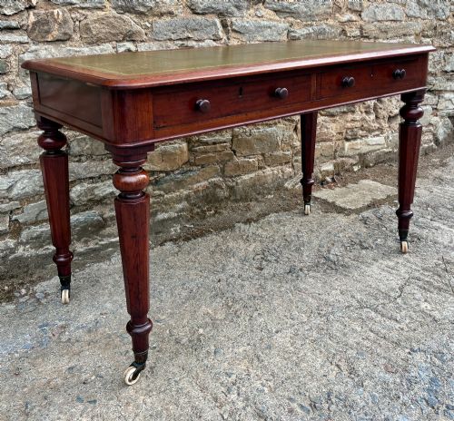 fig 2. A Victorian mahogany writing table featuring a green gilt-tooled leather writing surface, two drawers with turned knob handles, and turned tapered legs resting on ceramic castors.